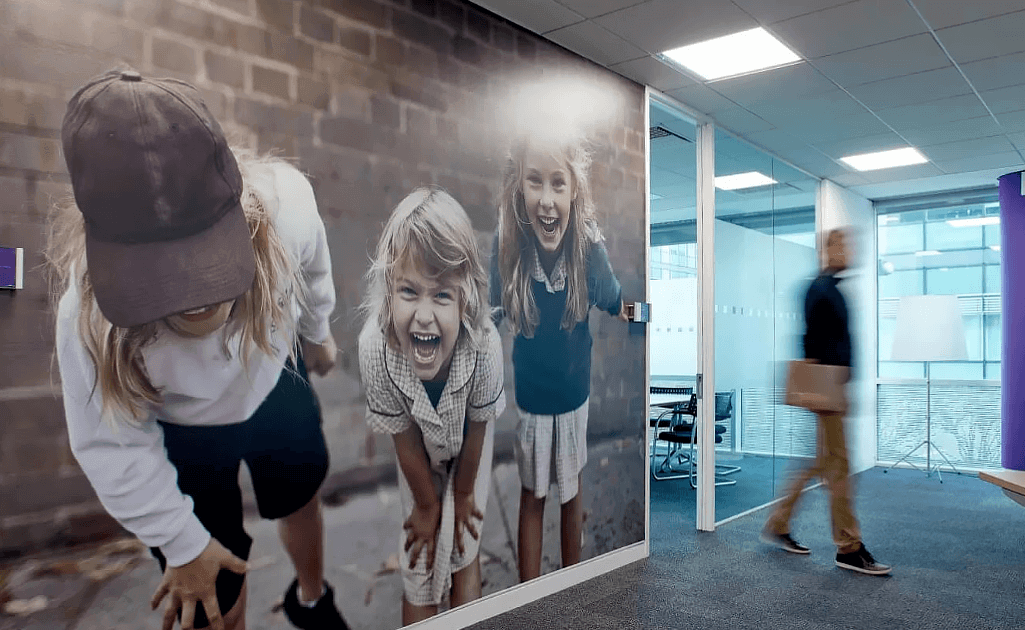 Aplicação de design gráfico em parede corporativa com imagem em grande escala de crianças sorrindo, transmitindo leveza e conexão emocional. A composição visual reforça a identidade da marca no ambiente, captando a atenção e comunicando com clareza por meio de impacto visual estratégico.